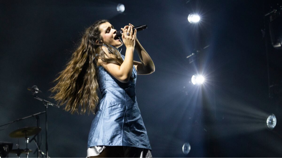 Cantante con micrófono en mano, vestido azul claro, actuando en un concierto con luces brillantes de fondo y parte de una batería visible.