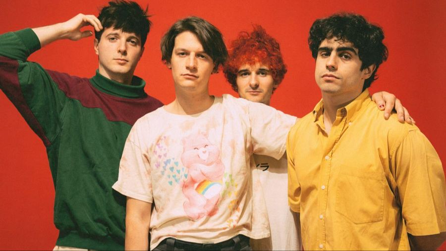 Cuatro jóvenes posan juntos sobre fondo rojo. Visten ropa informal: jersey bicolor, camiseta con osito, camiseta negra con letras y camisa mostaza.