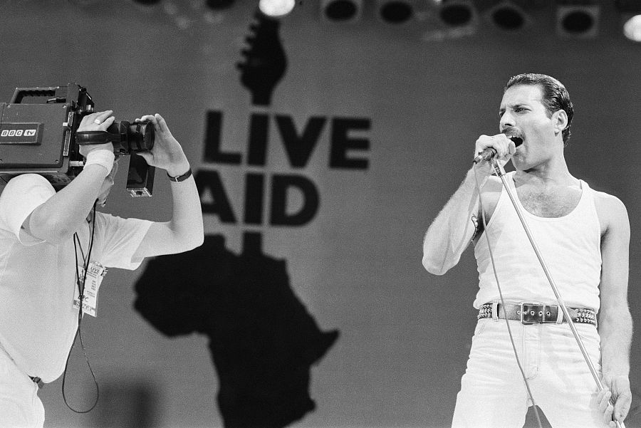 Freddie Mercury, en blanco y negro, canta con un micrófono en el Live Aid de 1985.  Viste camiseta y pantalones blancos; un cámara de la BBC lo graba.