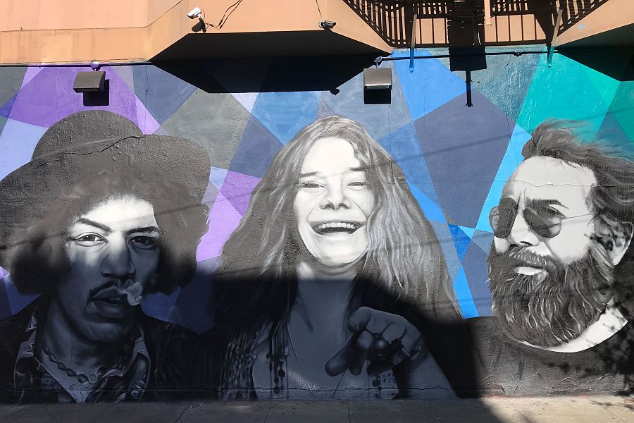Mural de tres músicos: uno con sombrero, otro sonriendo y señalando, y el último con gafas de sol y barba. Fondo geométrico azul, morado y gris.