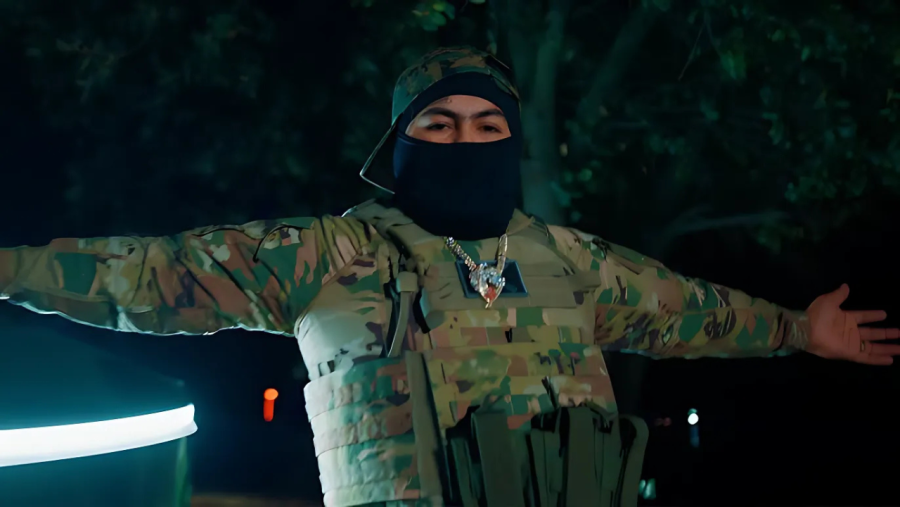 Artista musical en rodaje nocturno de videoclip.  Uniforme militar, chaleco antibalas, gorra y pasamontañas negros. Pose con brazos extendidos.