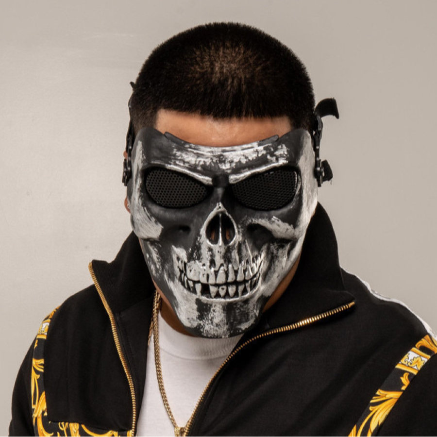 Rapero con máscara de calavera gris y negra, camiseta blanca y chaqueta negra con detalles dorados.  Malla negra cubre los ojos de la máscara.