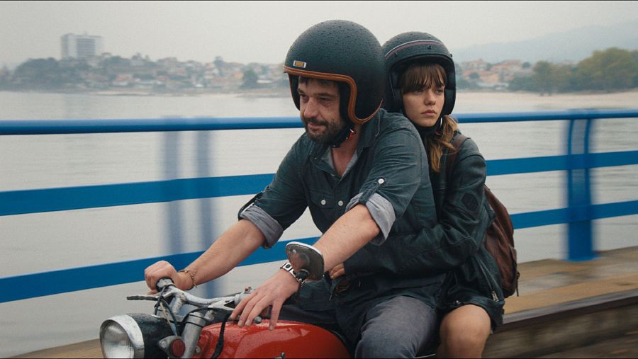 Pareja en moto roja cruzando un puente, ella abrazando al conductor. Cielo nublado y agua al fondo.