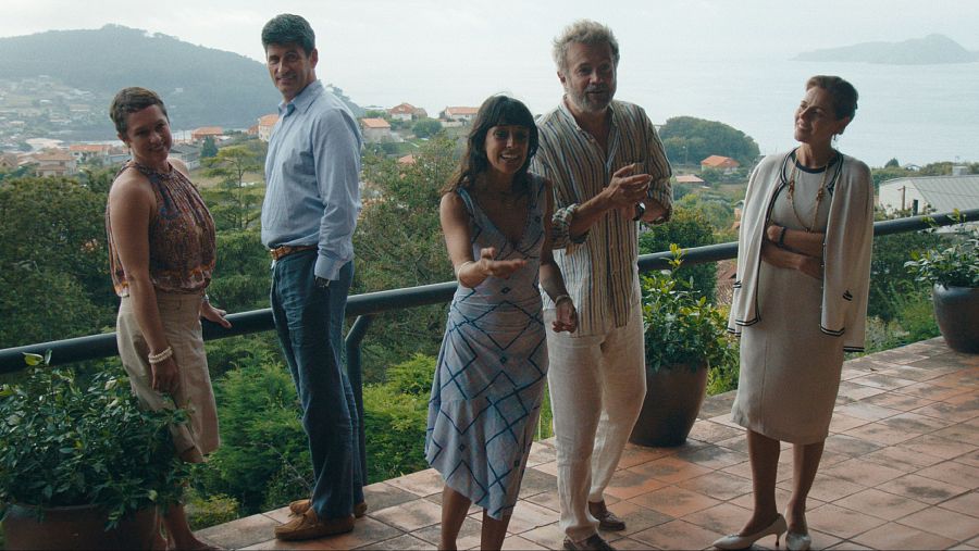 Cinco personas conversan en una terraza con vistas al mar. Una mujer con vestido azul claro gesticula; los demás la miran. Ambiente relajado y veraniego.