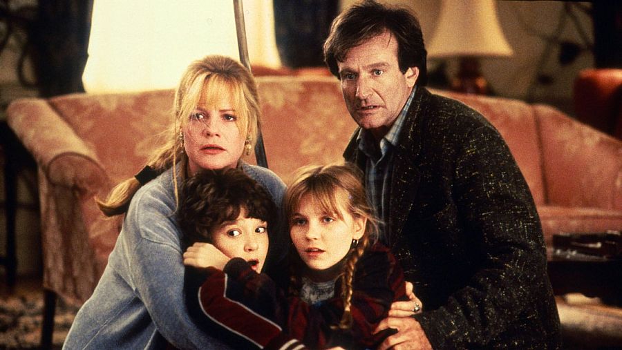 Robin Williams, Bonnie Hunt y dos niños, asustados y abrazados, en un salón con un sofá al fondo. Williams sostiene un objeto alargado.