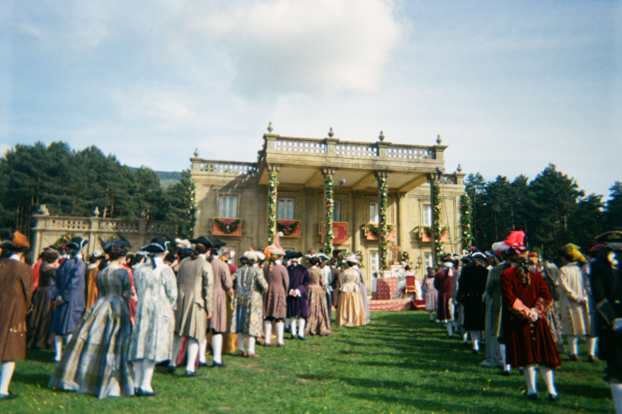 Multitud en traje de época (siglo XVIII) reunida en una campa ante un palacio decorado con guirnaldas y alfombra roja para una ceremonia o celebración.