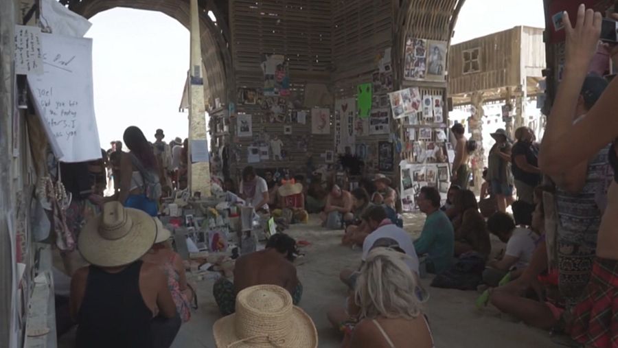 Interior de un espacio con paredes repletas de fotos y objetos personales. Personas sentadas en el suelo en actitud reflexiva. Ambiente de recogimiento y conmemoración.