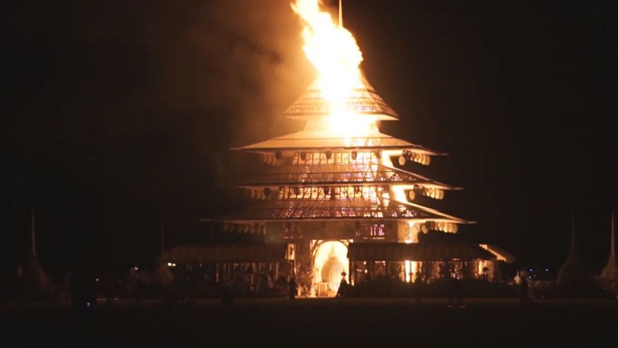 Estructura multi-nivel en llamas, iluminada por el fuego nocturno. Personas observan desde abajo.  Ambiente ritualístico.