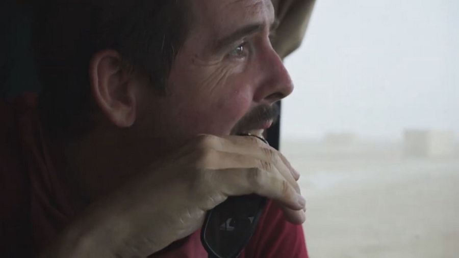 Hombre de perfil, con camisa roja, sostiene un dispositivo cerca de la boca y mira por una ventana a un paisaje exterior borroso con mal tiempo. Expresión seria y concentrada.