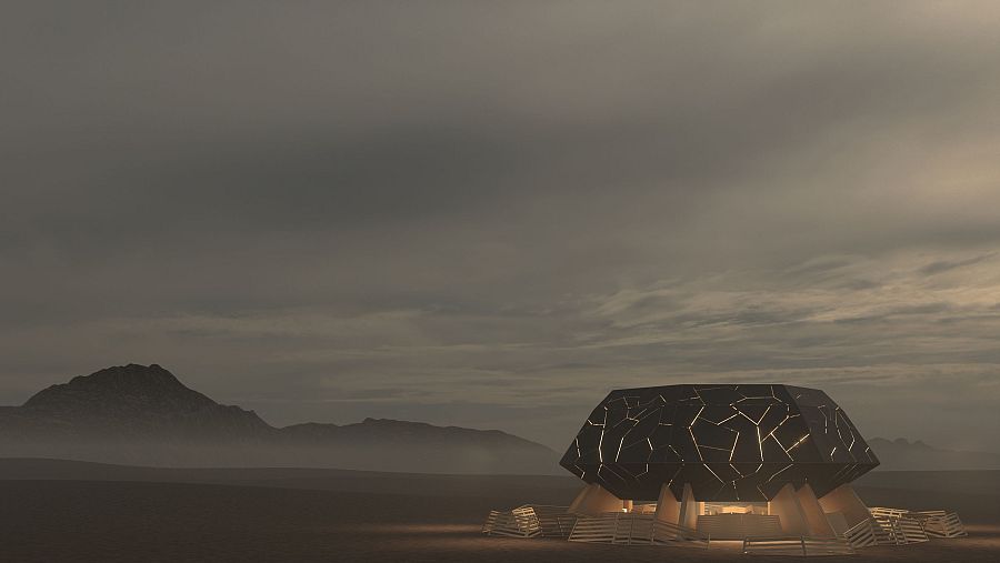 Estructura poligonal oscura con múltiples entradas, en un paisaje desértico con cielo nublado y montañas al fondo.  Rodeada por una valla de madera.