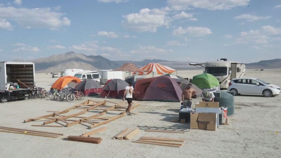Estructura de madera en construcción en un desierto, con voluntarios trabajando, tiendas de campaña, vehículos y materiales. Cielo parcialmente nublado.