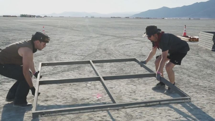 Trabajadores en un desierto montan una estructura metálica cuadrada.  Usan ropa informal y guantes.  Se ven montañas y materiales de construcción cerca.