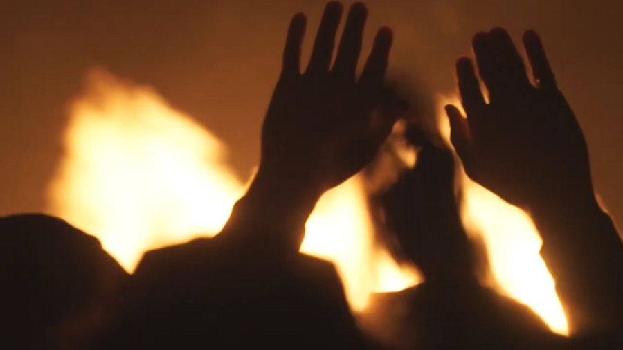 Siluetas oscuras alrededor de una fogata, con las manos levantadas.  La iluminación intensa destaca los contornos de las figuras.