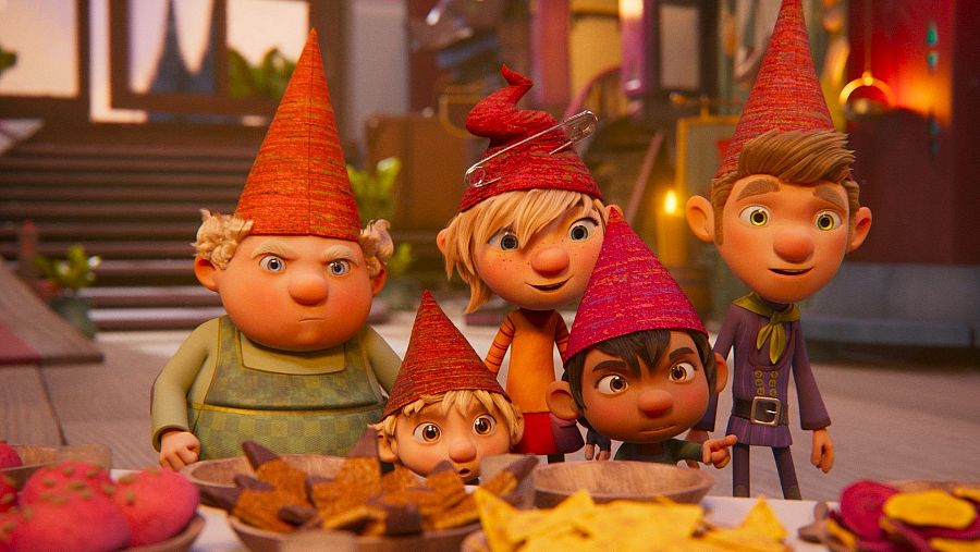 Escena animada con cinco elfos alrededor de una mesa con comida; gorros rojos y naranjas, expresiones variadas, ambiente acogedor.