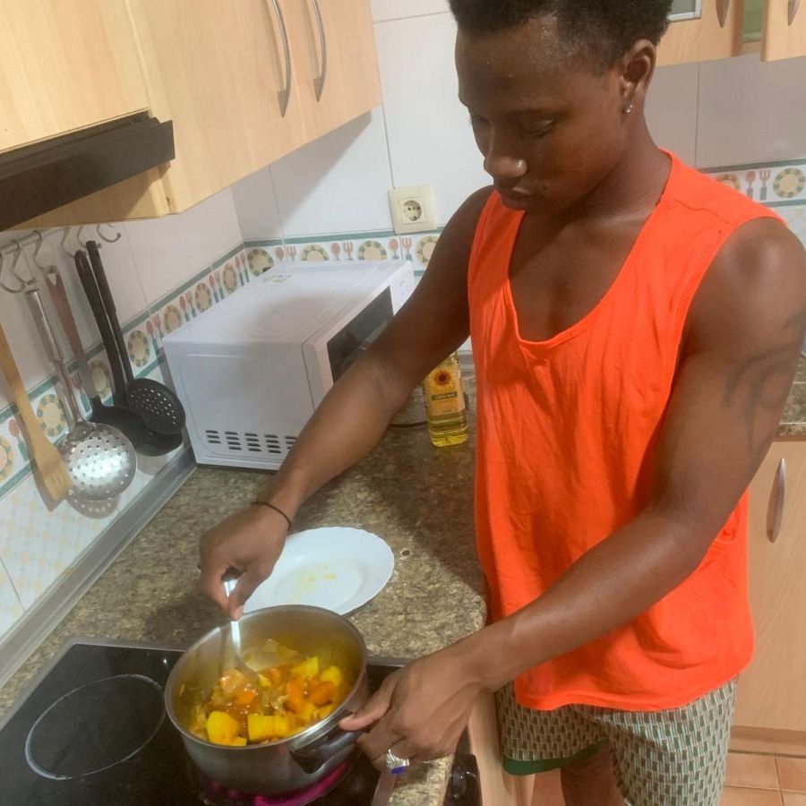 Joven cocinando un guiso en una cocina sencilla.  Usa una camiseta naranja y pantalones cortos.  Se observa una vitrocerámica, una olla y utensilios de cocina.