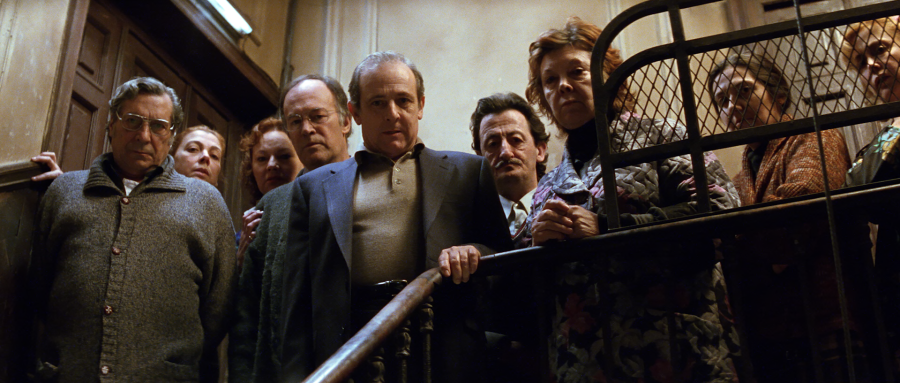 Escena de suspense en la escalera de un edificio antiguo. Varios personajes, con expresiones de sorpresa e inquietud, se encuentran en un ambiente de misterio.