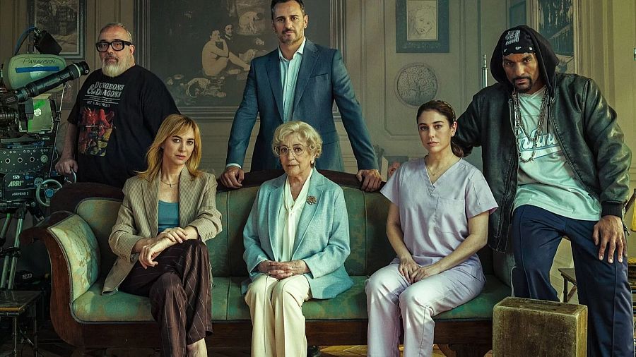 Álex de la Iglesia y elenco de 'La cuidadora' en foto promocional. Tres mujeres en sofá, dos hombres de pie, una cámara de cine visible. Ambiente clásico.