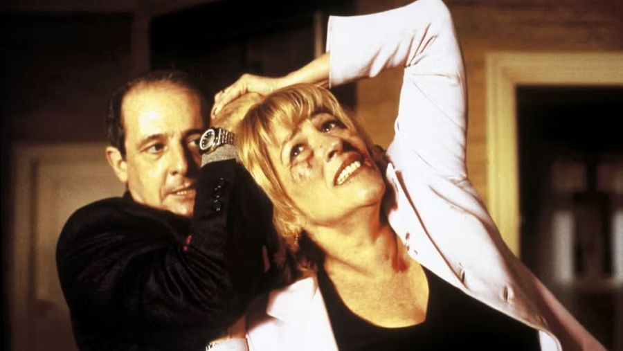 Escena de violencia en una película: un hombre sujeta a una mujer por el cuello, ella muestra signos de dolor y heridas. Iluminación tenue.