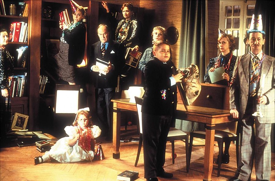 Escena festiva en una habitación desordenada con libros, niños jugando con serpentinas y adultos buscando entre libros.  Ambiente cálido y sugerente de un descubrimiento.