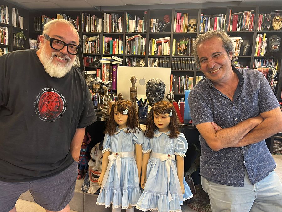 Entrevista a Álex de la Iglesia y Jose Fernández.  Él viste camiseta negra con una moneda y él camisa azul.  Fondo con estantería llena de objetos de colección.