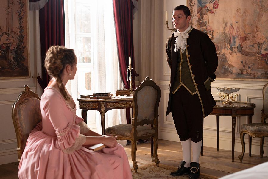Mujer con vestido rosa y hombre con traje marrón oscuro en una habitación elegante con muebles de madera y cortinas burdeos. Él parece entregarle una carta o libro.