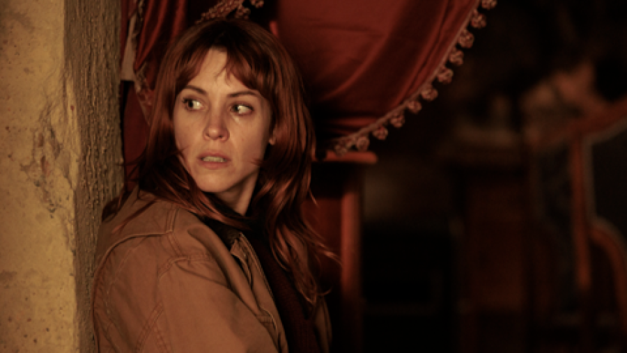 Mujer con pelo rojizo y chaqueta marrón, expresión de miedo en interior oscuro con cortina roja y pared rugosa. Atmósfera de suspense.