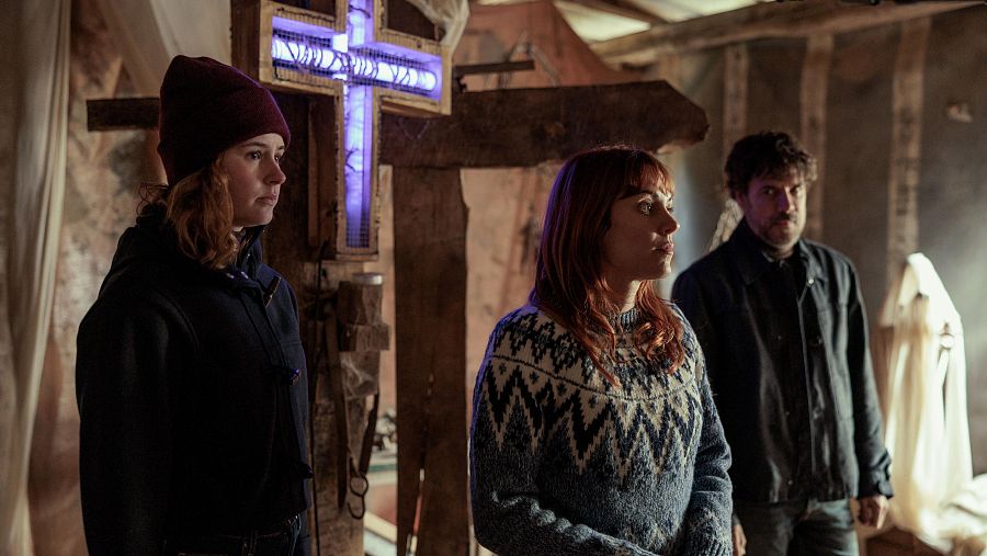 Tres personas, dos mujeres y un hombre, en una habitación rústica miran hacia un mismo punto. Una mujer lleva un gorro granate y la otra un jersey con motivos geométricos. Al fondo, una figura blanca cubierta.