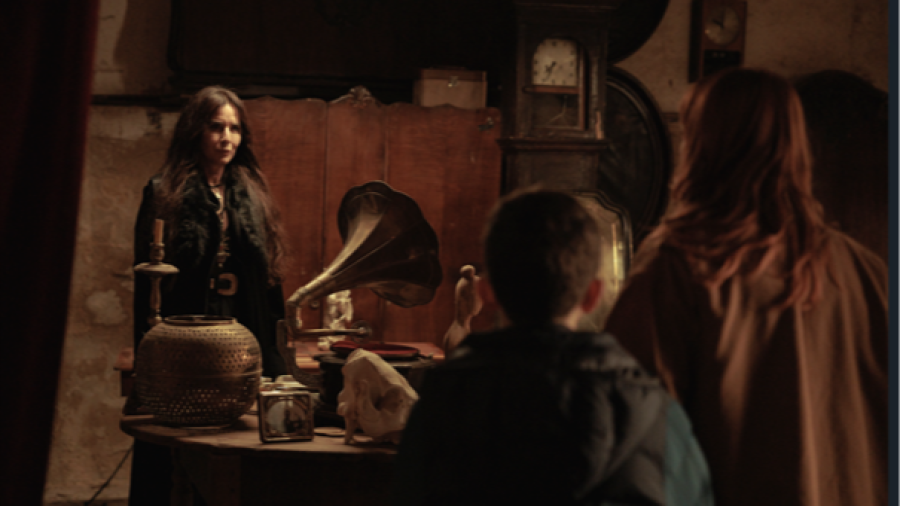 Tres personas en una habitación antigua con objetos como un gramófono y un candelabro. Una mujer con cabello oscuro, un niño y una mujer pelirroja observan la escena. Atmósfera misteriosa.