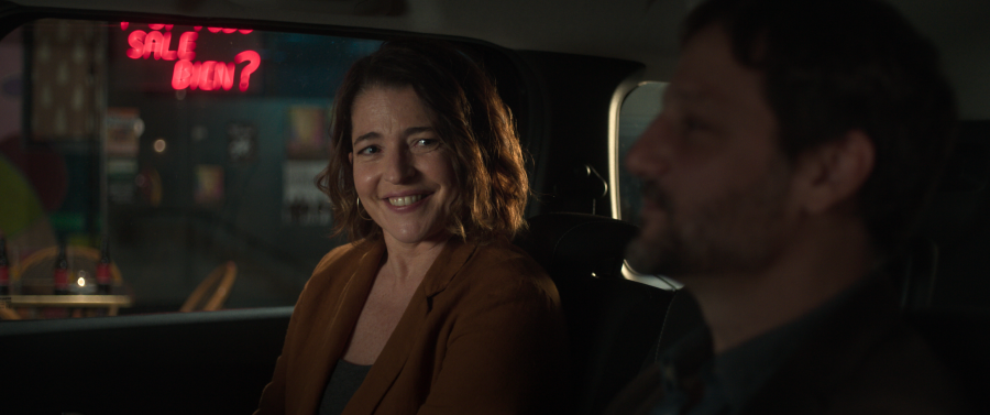 Interior de coche: mujer sonríe al conductor.  Letrero de neón rojo visible por la ventana. Ambiente relajado.