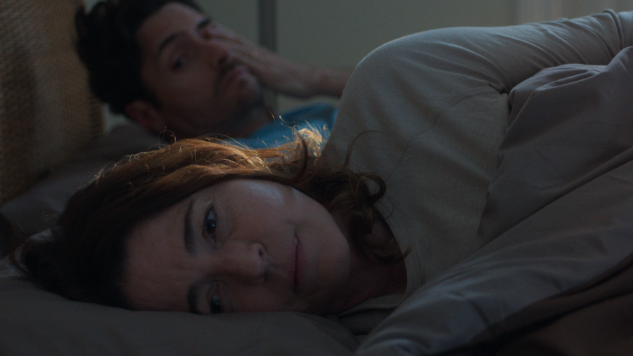 Escena nocturna en cama: mujer con cabello castaño rojizo mira a cámara con expresión seria; hombre con cabello oscuro, recostado, mano en cara.  Tensión palpable.