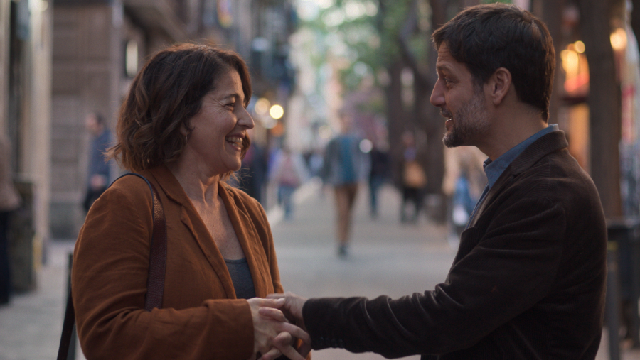 Pareja sonriendo y cogiéndose de las manos en una calle. Ella viste americana marrón y bolso; él, americana oscura y camisa azul. Ambiente urbano diurno.