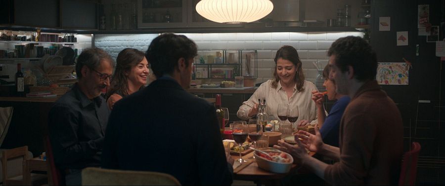 Cena familiar o entre amigos en una cocina, con siete personas alrededor de una mesa de madera. Ambiente cálido y acogedor, con azulejos blancos y pizarra con dibujos infantiles.