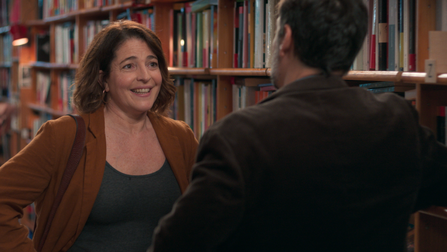 Escena de una librería: mujer con chaqueta marrón anaranjado y bolso, sonríe a cámara mientras conversa con un hombre de espaldas.