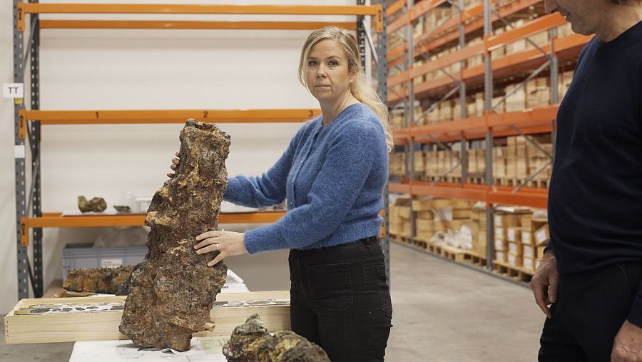 Geóloga muestra roca marrón oscura de fondo marino, rica en cobre y otros minerales, en almacén con estanterías naranjas.