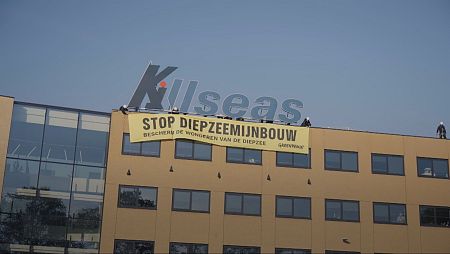 Protesta en el tejado de un edificio. Pancarta amarilla con mensajes en neerlandés contra la minería de aguas profundas y el logo de Greenpeace. Activistas presentes.