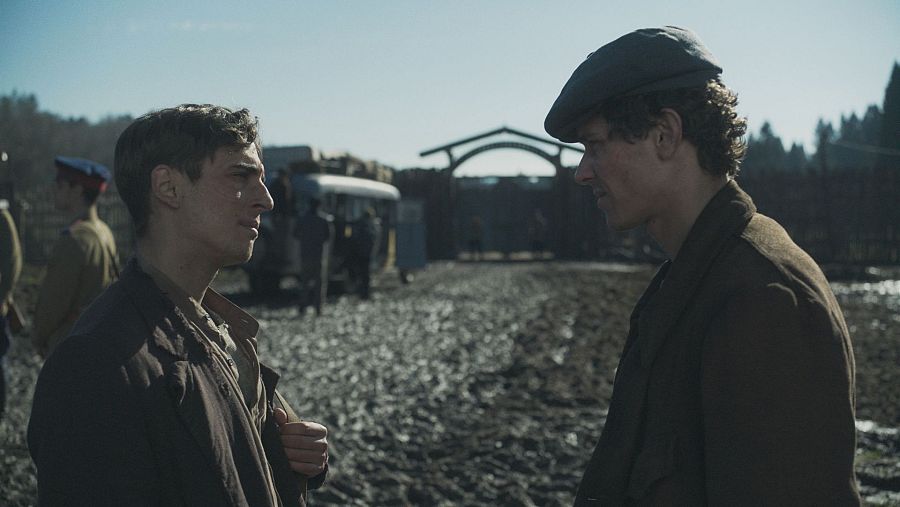 Dos jóvenes, uno con boina, se miran en un camino de tierra.  Fondo con estructura de madera, vehículo antiguo y posibles militares. Contexto histórico bélico o posguerra.