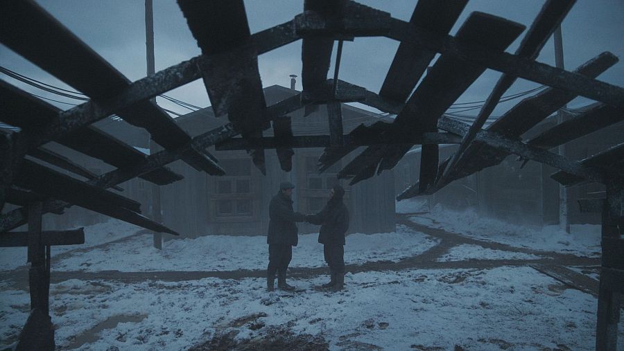 Dos hombres se dan la mano en un paisaje nevado, con ruinas de madera al fondo.  Escena de reconciliación en atmósfera fría y sombría.