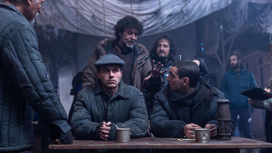 Director dirigiendo a dos actores sentados a una mesa con una lámpara de aceite; ambiente oscuro y rústico, con otros miembros del equipo de rodaje presentes.