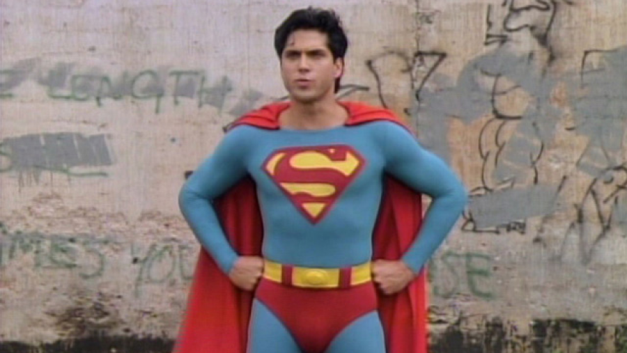 Superboy, interpretado por John Haymes Newton, posa con su traje azul, capa roja y símbolo 'S'. Fondo con grafitis.