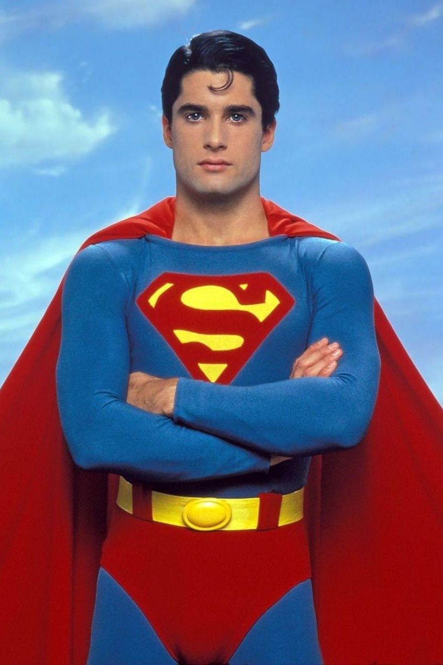 Superboy, con traje azul y capa roja, posa con los brazos cruzados sobre un fondo de cielo azul. El icónico símbolo de Superman destaca en su pecho.