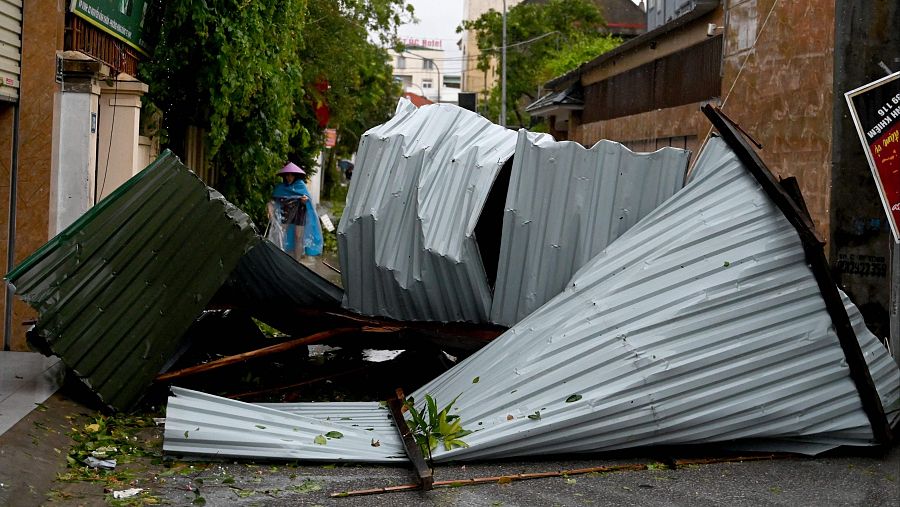 Escombros metálicos doblados y retorcidos en una calle tras un tifón. Una persona con chubasquero azul limpia entre los restos. Daños menores en el fondo.