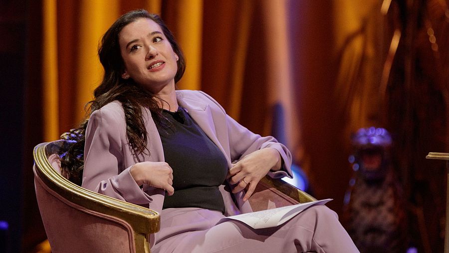 Victoria Martín, en un sillón rosa, viste traje lila y camiseta negra; su expresión indica que habla o escucha. Fondo: posible plató de televisión.