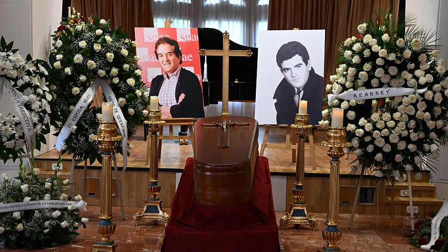 Féretro cerrado con cruz, rodeado de coronas de flores y candelabros con velas.  Dos fotografías del difunto al fondo, una a color y otra en blanco y negro.