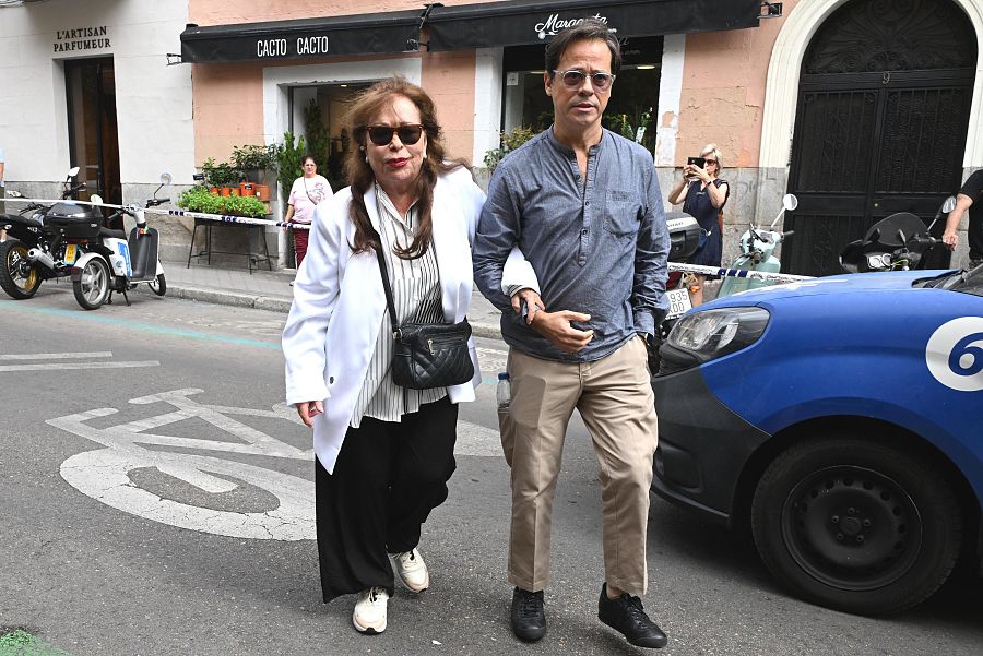 Massiel llega a una capilla ardiente acompañada de un hombre; ambos caminan por una calle con presencia policial y varios viandantes observan.