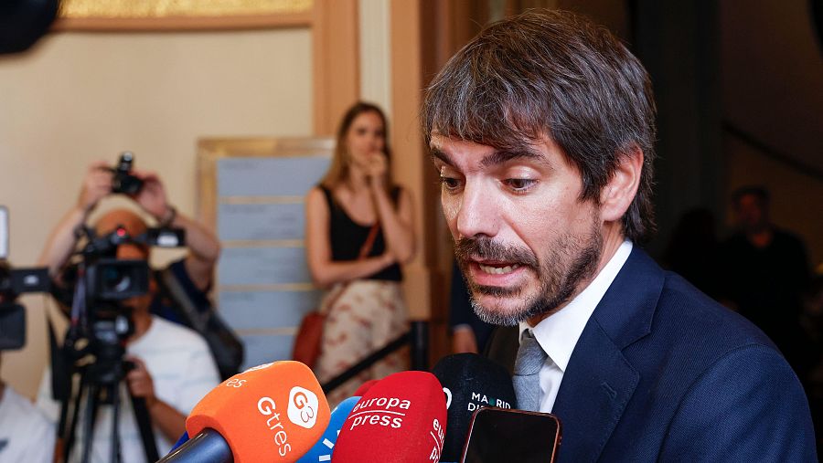 Eurodiputado hablando ante micrófonos de prensa en un evento público.  Se observa a una mujer en segundo plano.