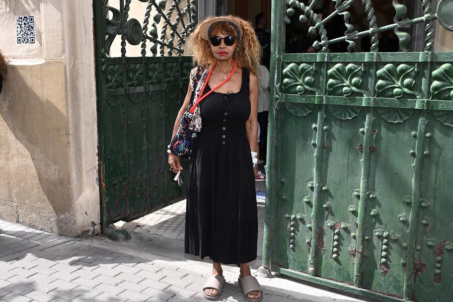 Mujer con vestido negro, sombrero y gafas de sol, frente a una puerta verde ornamentada de metal.  Detalles de bolso multicolor y sandalias grises.