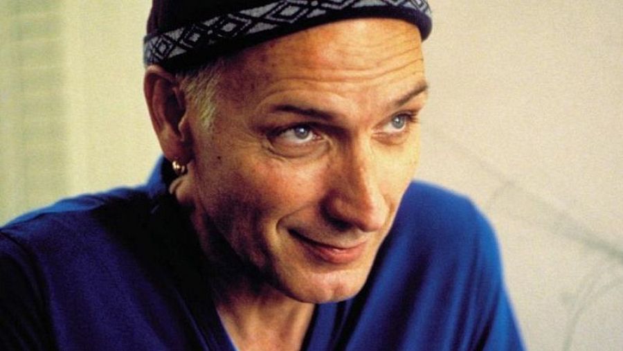 Primer plano de Eusebio Poncela; gorro de lana oscuro con diseño geométrico, prenda azul, pelo canoso corto, expresión seria y leve sonrisa, pendiente en oreja izquierda.