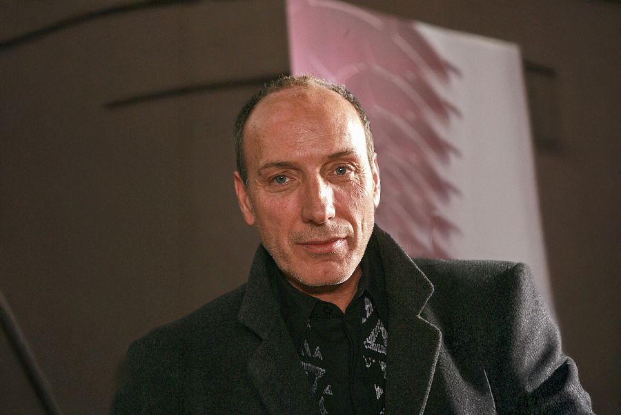 Retrato de Eusebio Poncela, de mediana edad, con cabello entrecano y una expresión serena. Lleva una chaqueta oscura sobre camisa negra con diseño geométrico. Fondo oscuro.