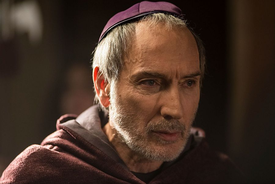 Actor con túnica granate y bonete morado, interpretando un personaje serio y reflexivo.