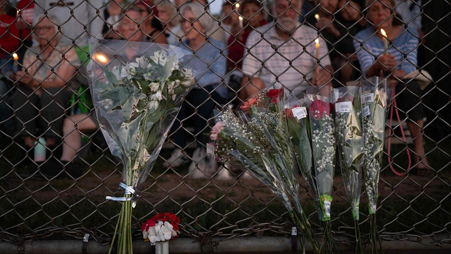 Primer plano de varios ramos de flores en plástico transparente apoyados en una valla metálica; al fondo, personas con velas encendidas en un acto conmemorativo.